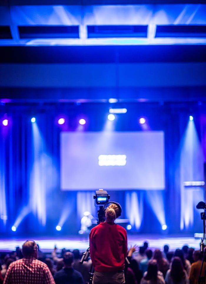 Cameraman capturing a live event on stage with colorful lights and blurred audience.