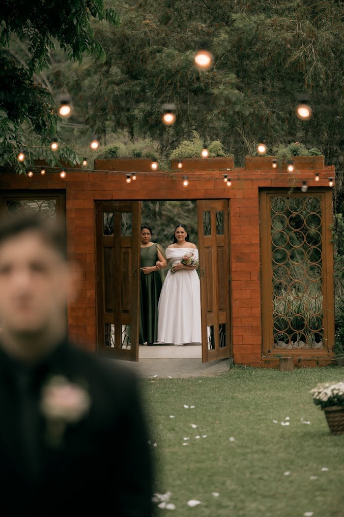 Bride and bridesmaid walking outdoors towards wedding ceremony in garden setting.